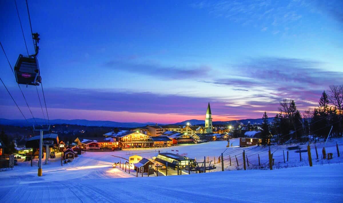 A vibrant ski resort village glows under a twilight sky, with illuminated buildings, snow-covered slopes, and a ski lift gondola in the foreground, set against a backdrop of distant mountains.