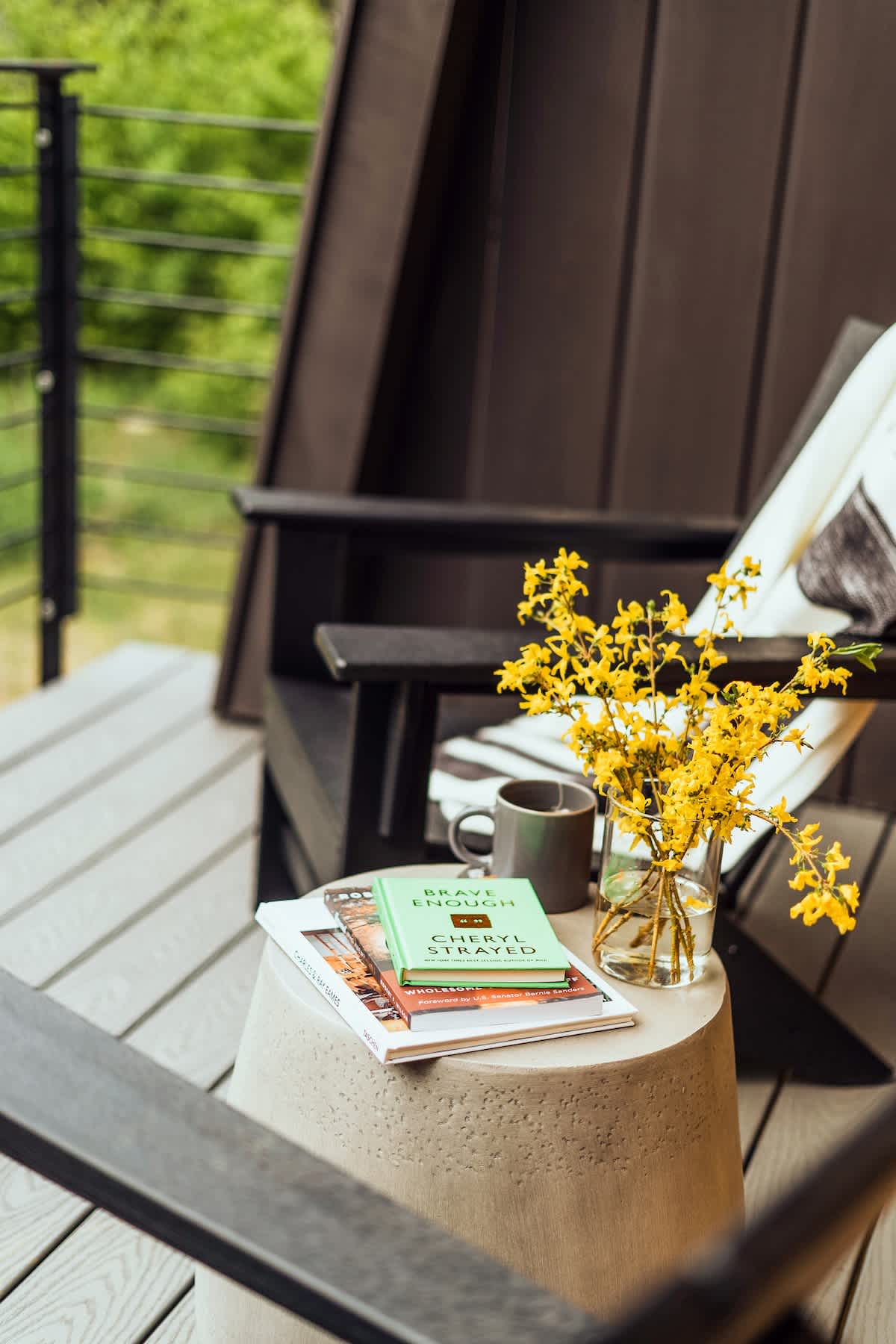 A cozy outdoor balcony scene with dark Adirondack chairs and a concrete side table holding books, a gray mug, and a vase of bright yellow flowers. A dark railing and green foliage are visible in the background.