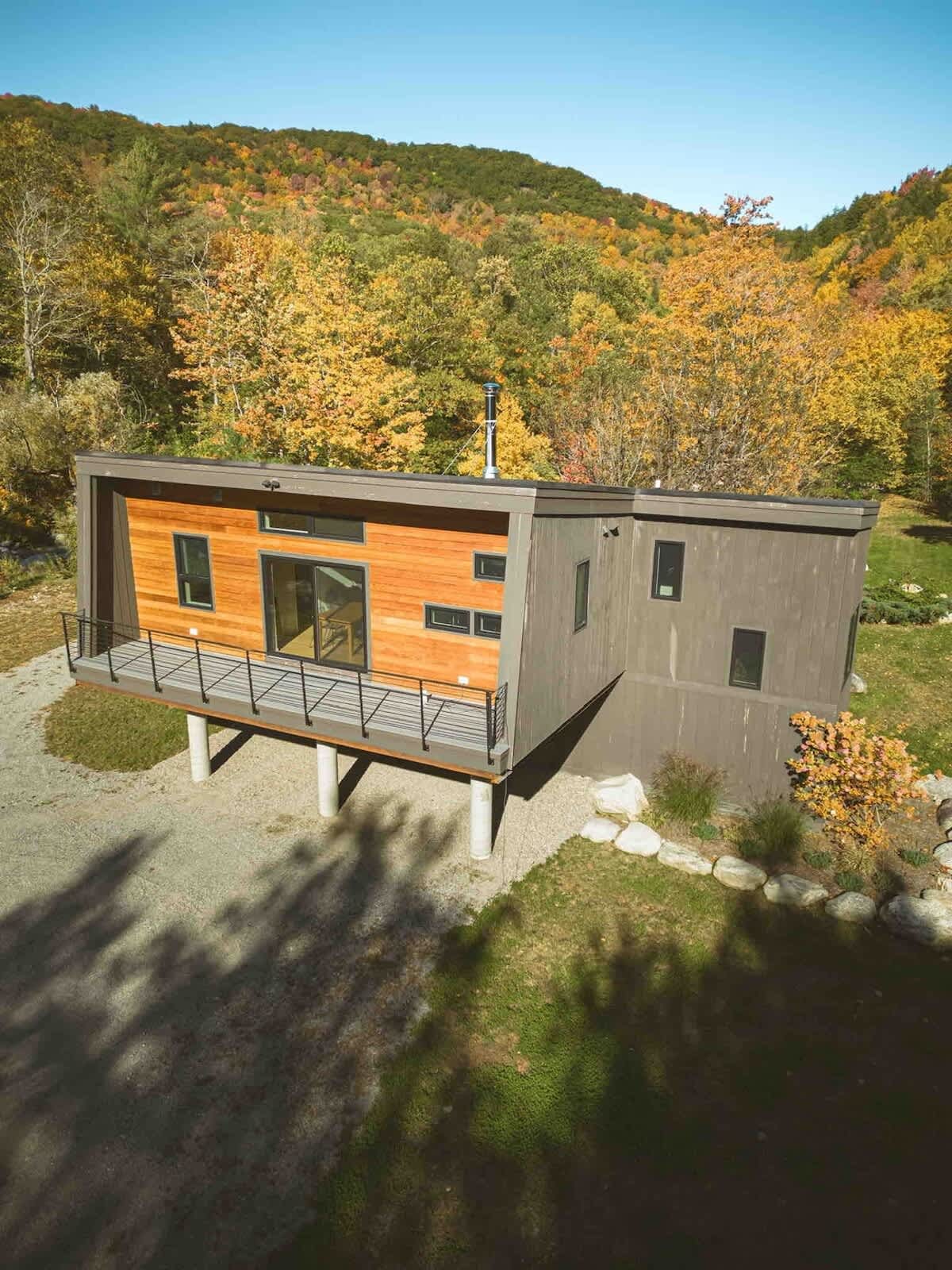 Modern elevated house with a mix of wood and dark gray siding, featuring a front balcony with a black railing. The house is supported by concrete pillars, with a gravel driveway below, and surrounded by autumn trees.
