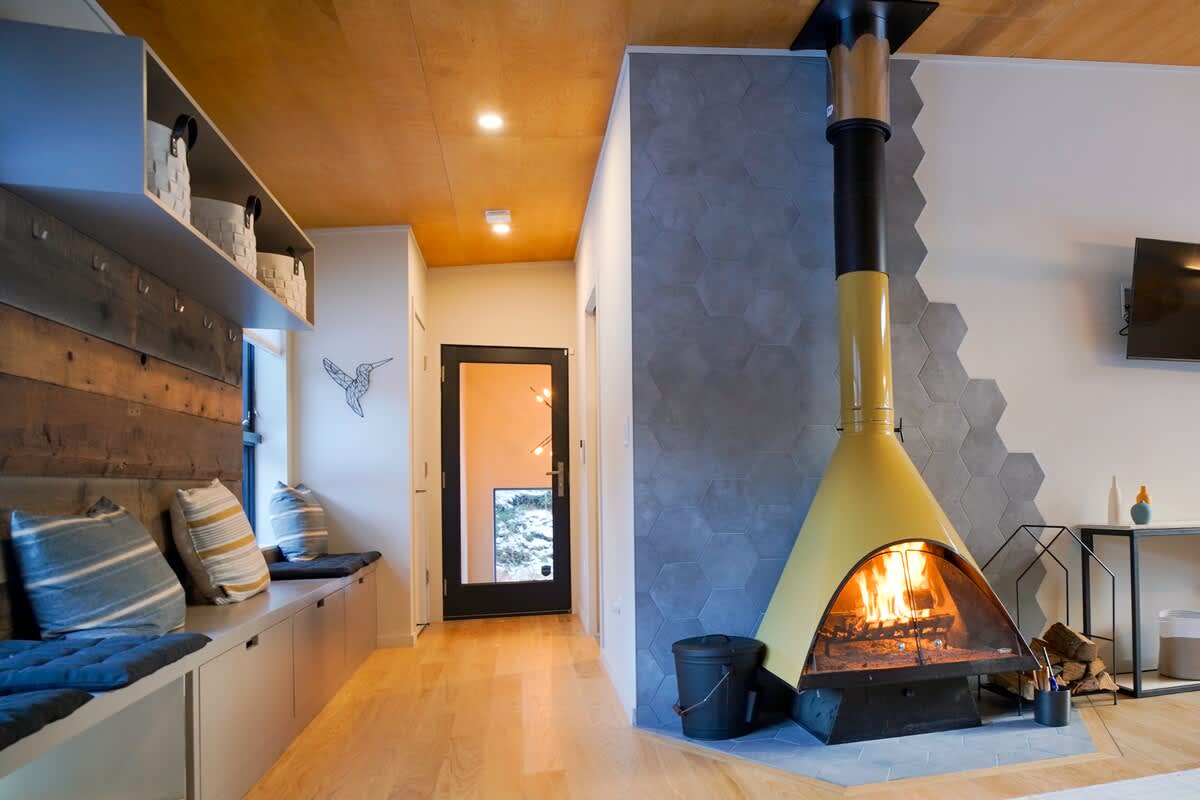 A bright foyer area features a long built-in window seat with storage, a rustic wooden accent wall, and a warm wood-paneled ceiling. A yellow freestanding fireplace with a hexagonal tile surround adds a focal point, leading to a black door with glass panels.
