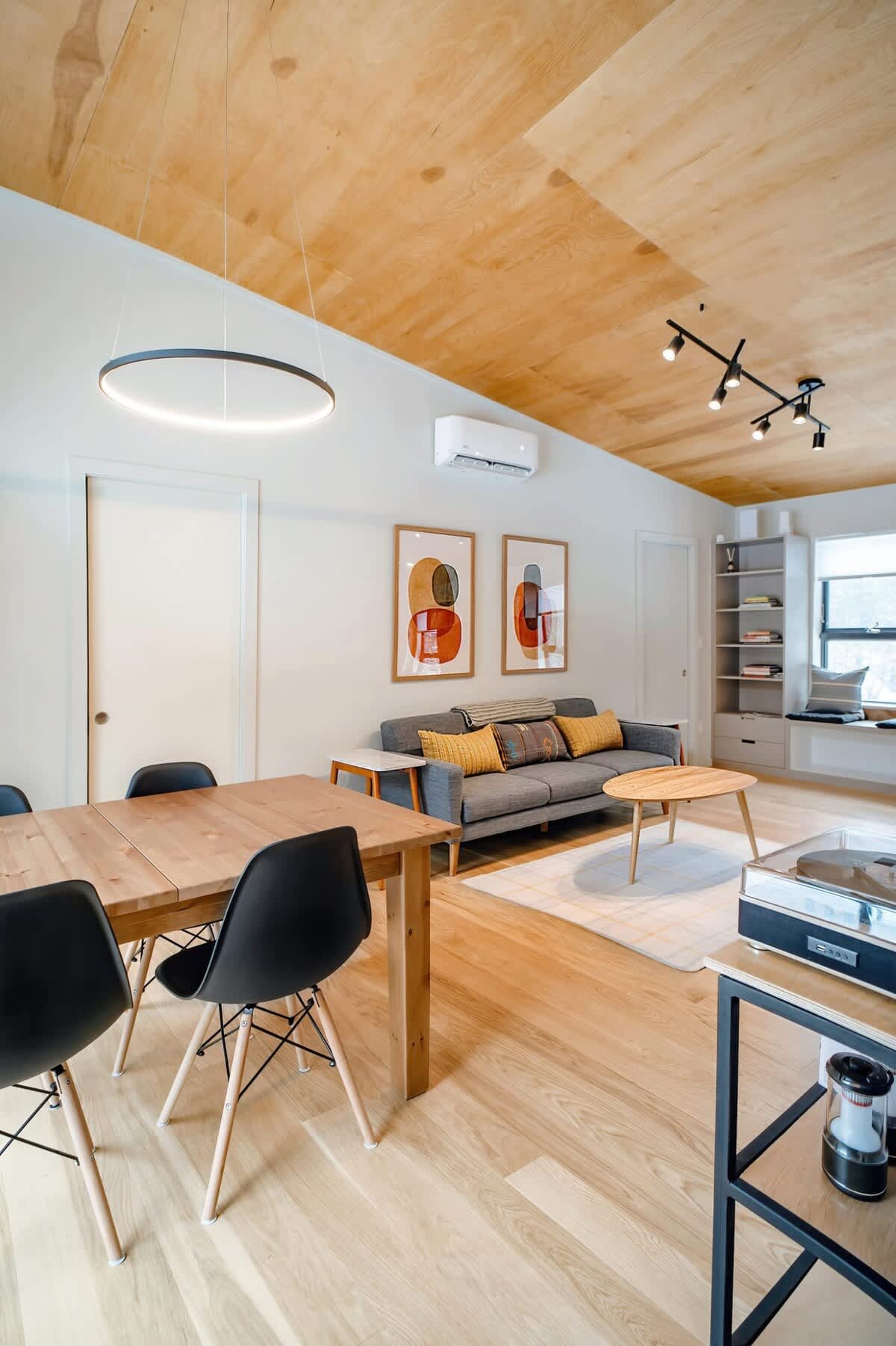 Modern open-concept living and dining space with light wood floors and a matching wood-paneled ceiling. A light gray sofa with colorful throw pillows faces a coffee table, while a wooden dining table with black chairs sits under a circular pendant light.