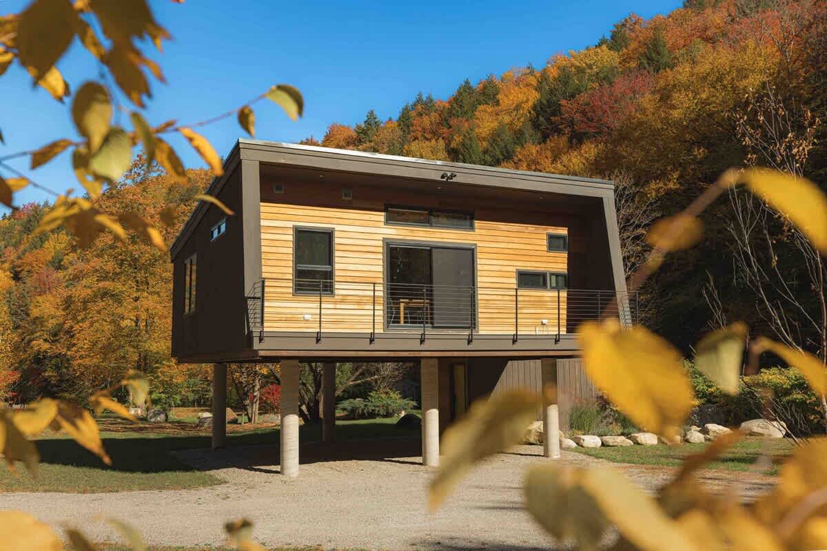 Modern elevated house with wood and dark gray siding, featuring a front balcony with a railing, set against a vibrant autumn forest backdrop. Yellow leaves are blurred in the foreground.