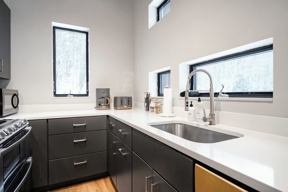Contemporary kitchen with dark gray flat-panel cabinetry, white quartz countertops, and stainless steel appliances. Multiple windows, including high clerestory windows, provide natural light, and a gooseneck faucet is visible over the sink.