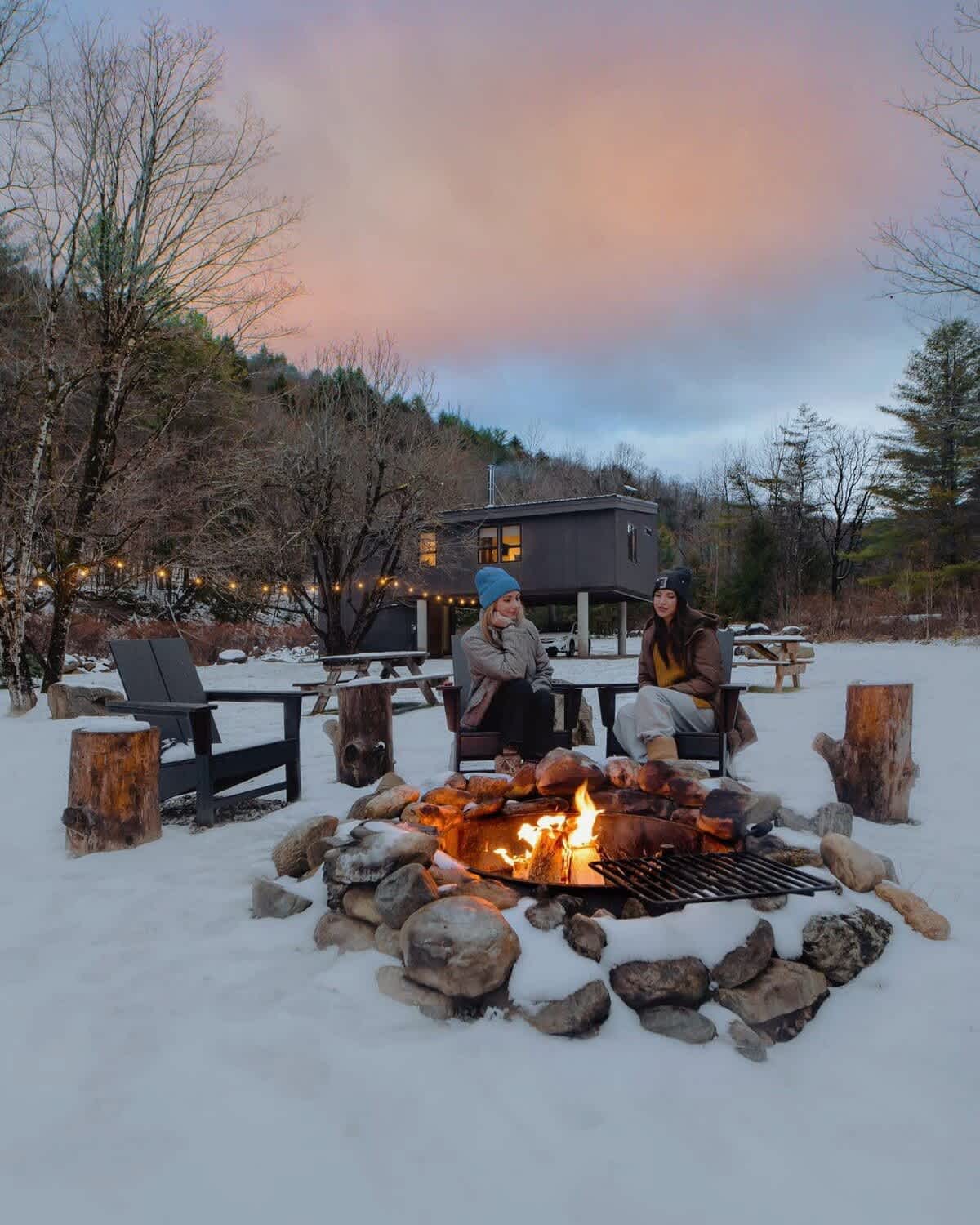 Two people sit around a stone fire pit with a grill grate, enjoying a fire in a snowy outdoor setting at dusk. The property's modern cabin structure is visible in the background, surrounded by trees and string lights.