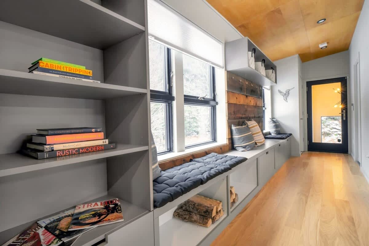 A long hallway or mudroom area with built-in gray shelving and a window seat featuring dark cushions. The window seat offers a view of the outdoors, and stacked firewood is stored beneath it, alongside books on the shelves.