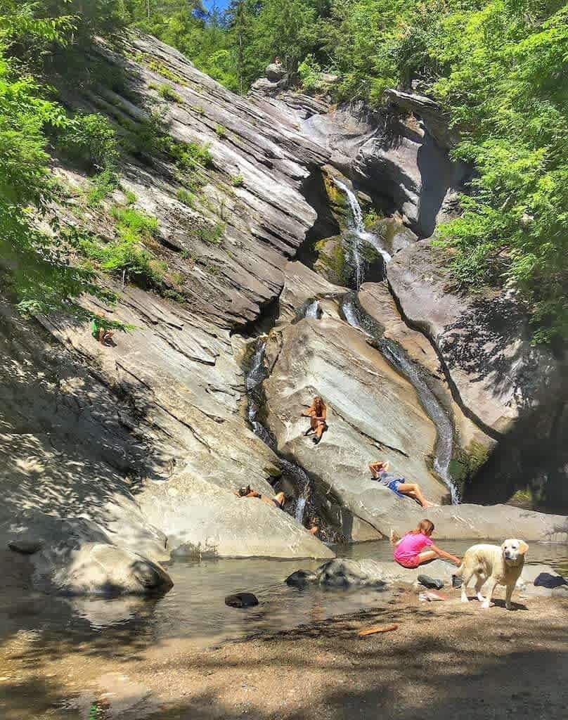 People and a dog enjoy a natural rock slide and shallow pool at the base of a cascading waterfall, surrounded by lush green foliage on a sunny day. The rock formations are smooth and worn by water.
