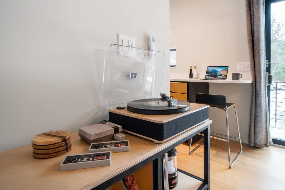 A dedicated entertainment corner featuring a record player and a vintage gaming console on a light wood and metal console table. In the background, a small breakfast bar with a laptop and stool is visible, leading to an outdoor view.