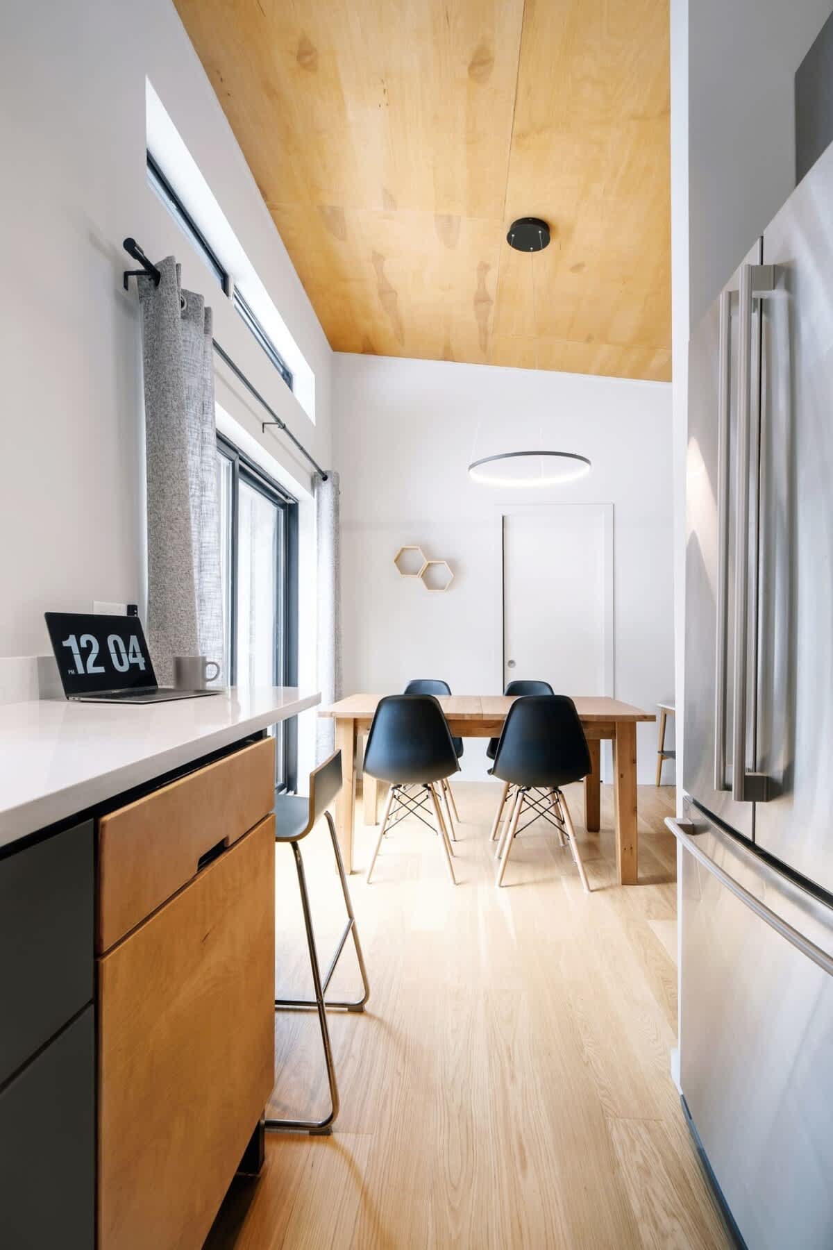 View from the kitchen showing a modern dining area with a light wood table and black chairs, illuminated by a minimalist circular pendant light. The kitchen counter with a laptop and stool is visible in the foreground, and a stainless steel refrigerator stands to the right.