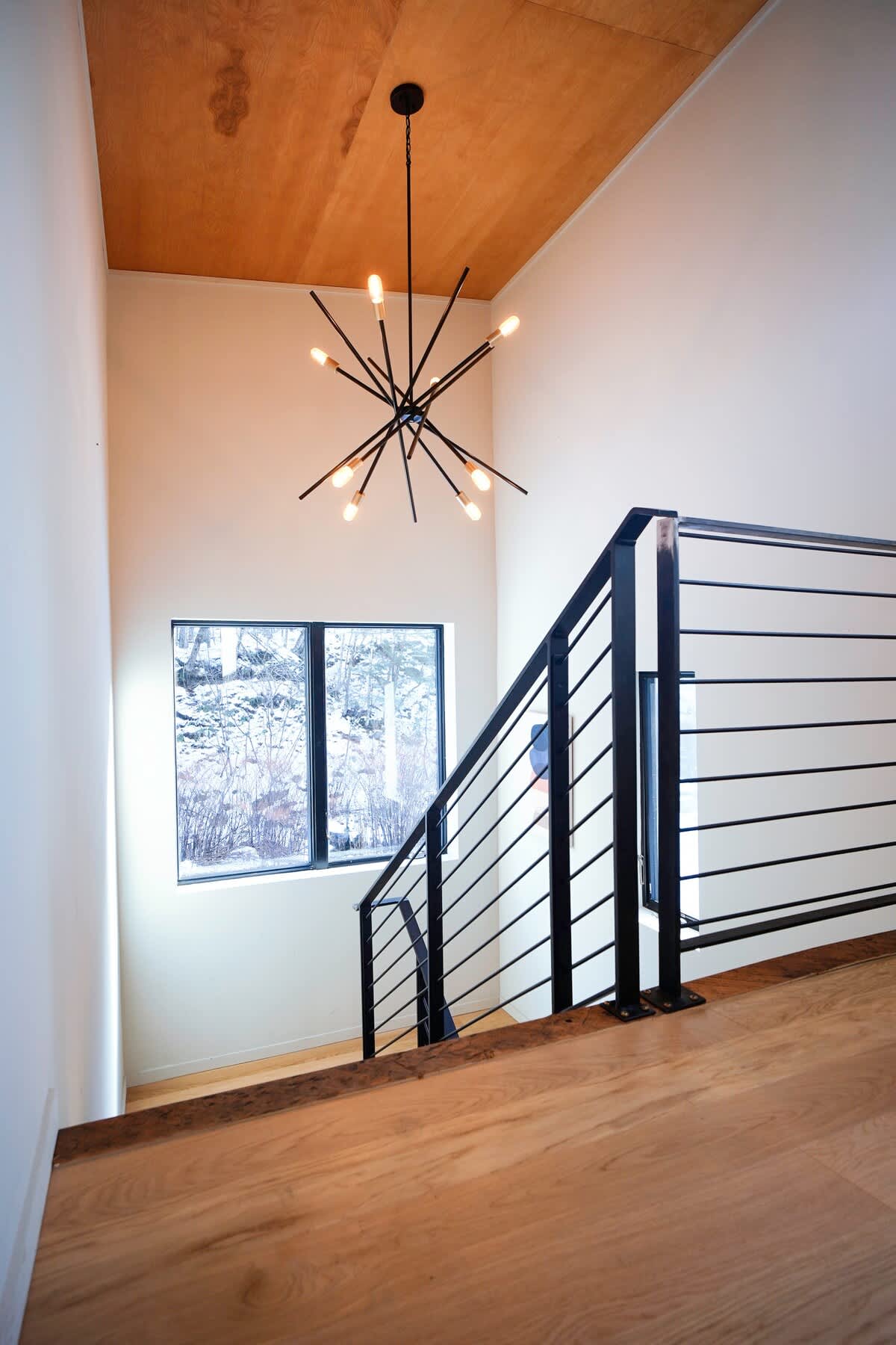 Modern staircase with light wood treads and a black metal railing featuring horizontal bars. A contemporary sputnik-style chandelier hangs from the high ceiling, and a large window offers a view of the snowy outdoors.