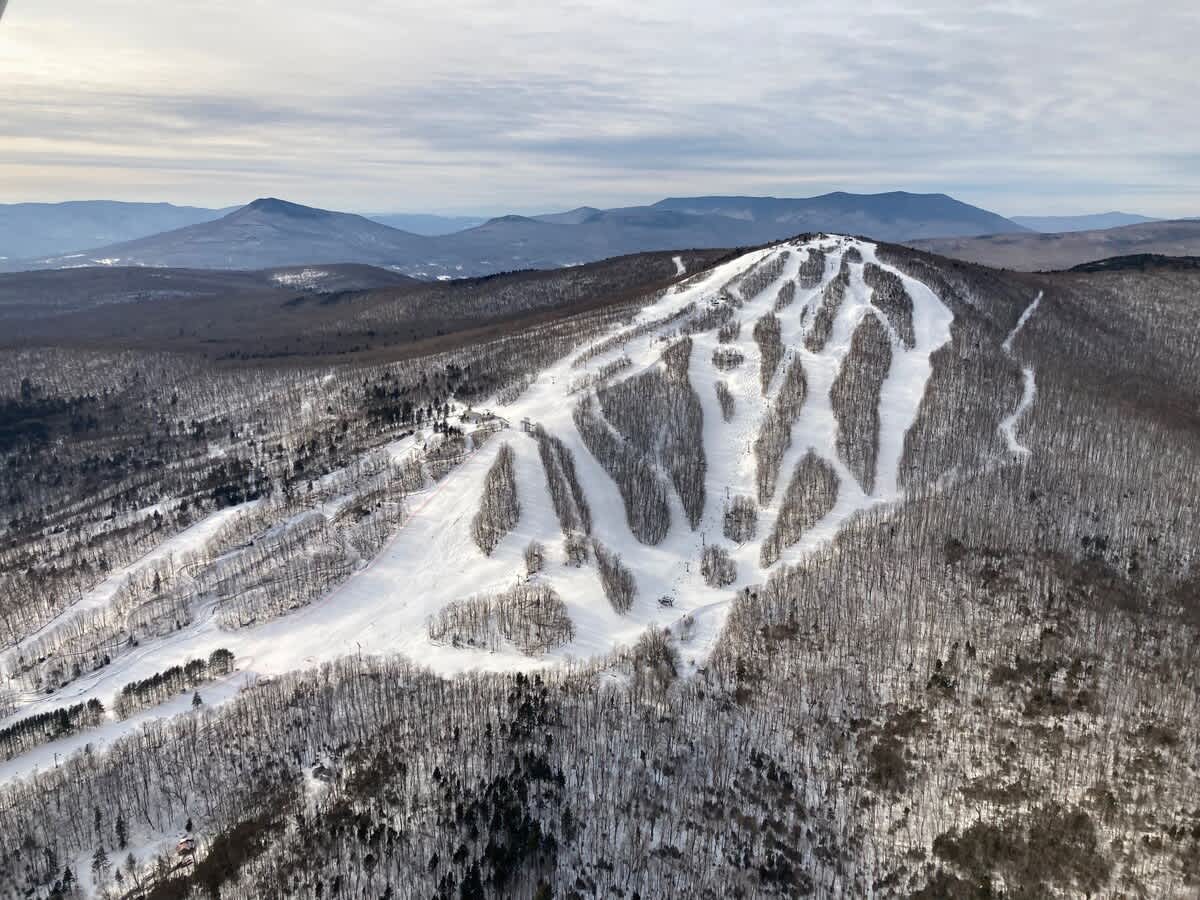 An expansive aerial view showcases a snow-covered ski mountain with numerous groomed runs winding through dense, frosted evergreen forests, under a cloudy winter sky.
