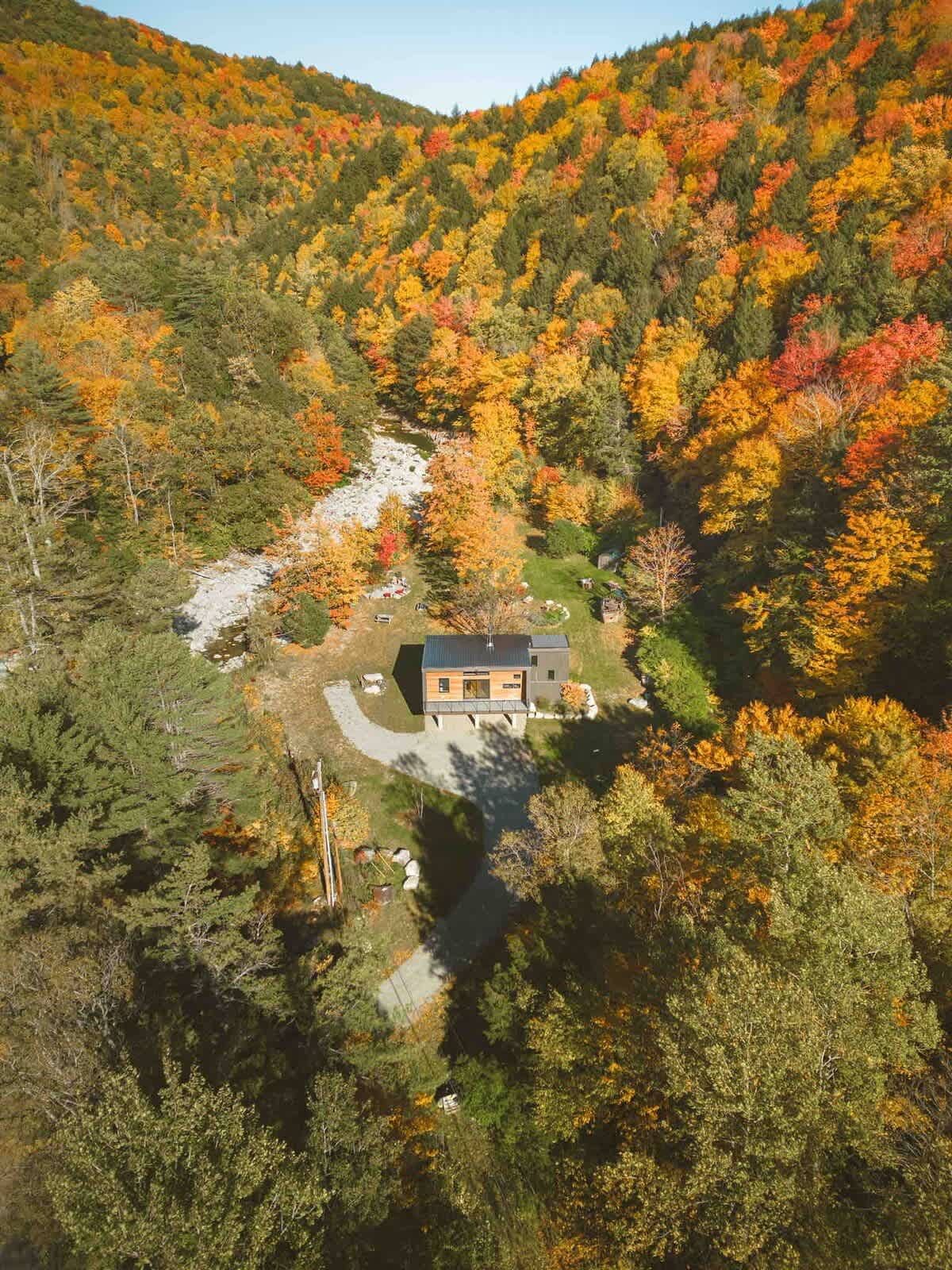 Aerial view of a modern house with a dark roof and wood siding, nestled in a valley surrounded by a winding river and dense forest with vibrant autumn foliage. A gravel driveway leads to the house.