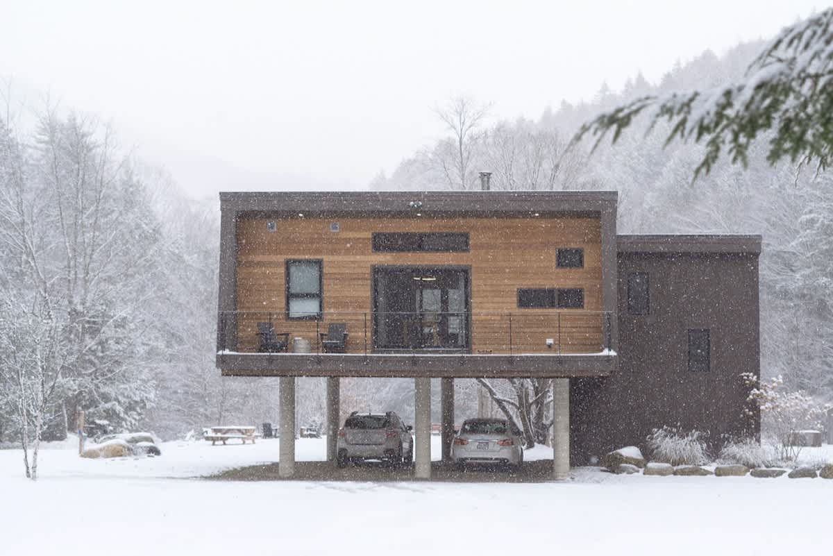 Modern elevated house with wood and dark gray siding, seen during a snowfall, with two cars parked underneath its elevated structure. The surrounding landscape is covered in fresh white snow, with snow-dusted trees in the background.