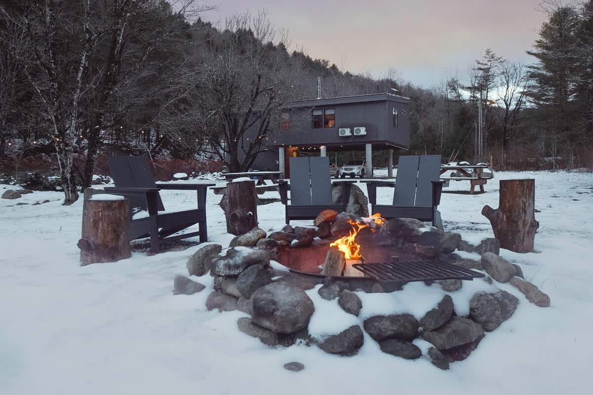 Outdoor fire pit area surrounded by rocks and tree stump seats, with Adirondack chairs in a snowy landscape. The house is visible in the background.