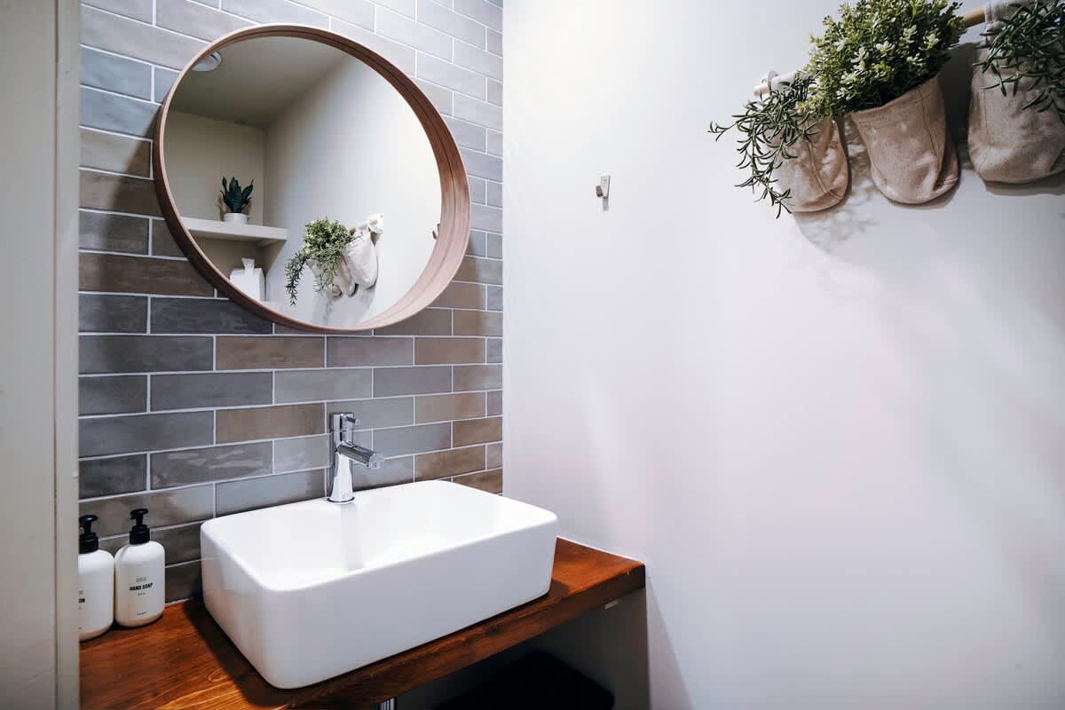 A modern bathroom vanity features a rectangular vessel sink on a wooden countertop, complemented by a round wooden-framed mirror above. The accent wall is tiled with gray and brown subway tiles, and potted plants add a natural touch.