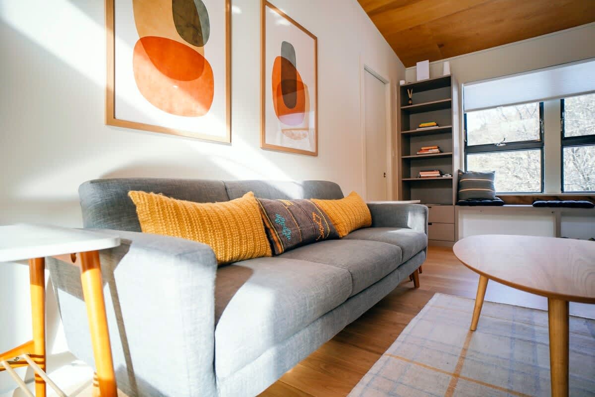 Close-up of a modern living room featuring a light gray sofa adorned with mustard yellow and patterned throw pillows. Two abstract art pieces hang above the sofa, and a light wood coffee table sits on a patterned rug.