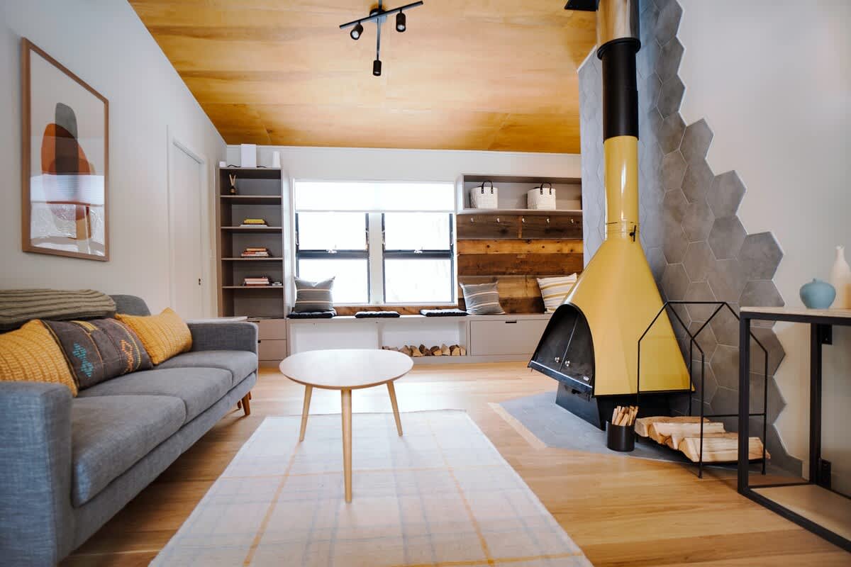 A modern living room features a striking yellow freestanding fireplace, a comfortable gray sofa with accent pillows, and a light wood coffee table on a plaid rug. The room boasts a warm wood-paneled ceiling and a built-in window seat with shelving.