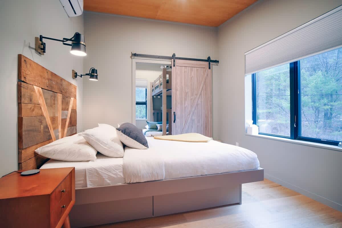 Another view of bedroom-1, showing a queen bed with a rustic wooden headboard, light wood floors, and a large window with blinds. A barn door leads to an adjacent room.