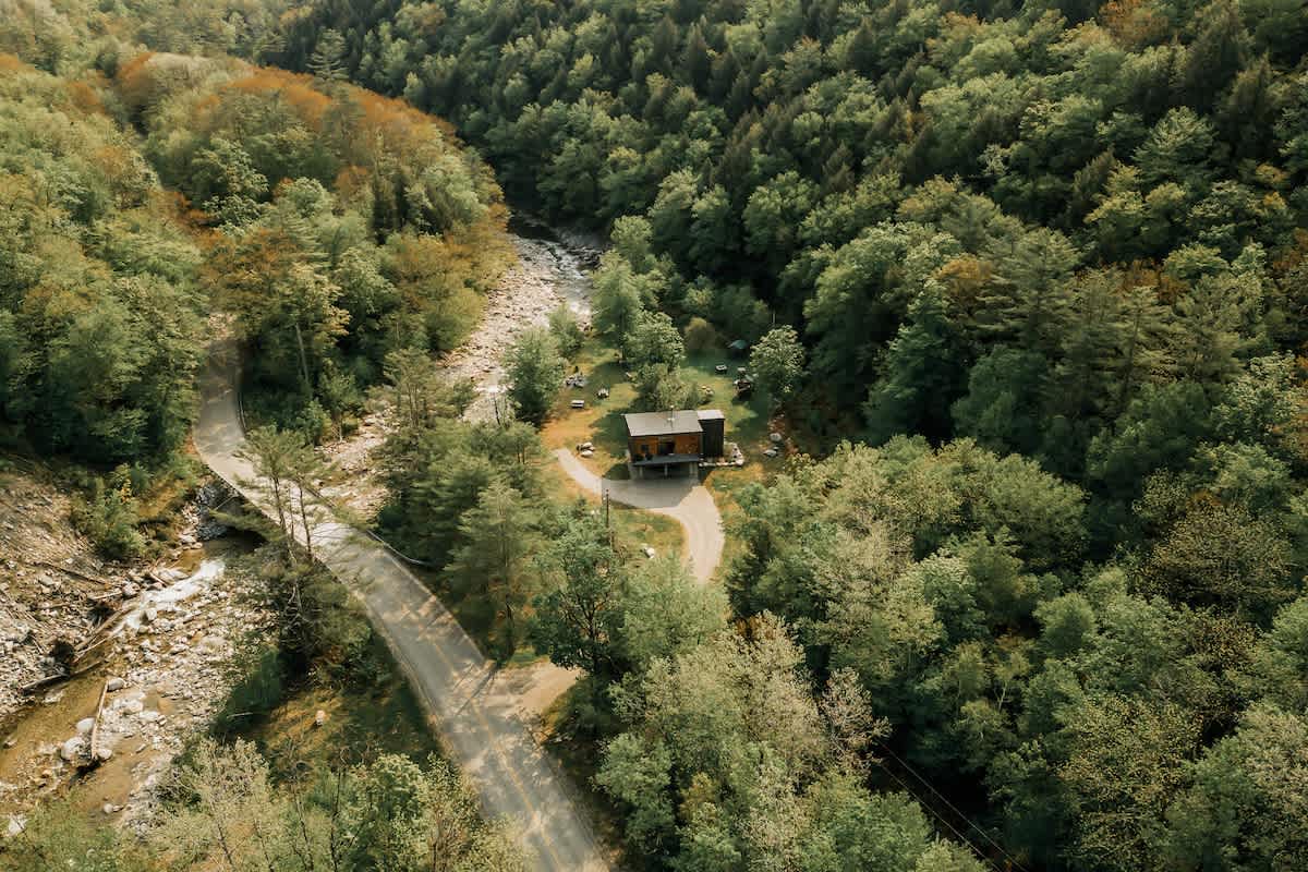 Aerial view of a modern house with a dark roof and wood siding, situated in a lush green valley with a winding river and a gravel road. The surrounding forest shows early signs of autumn color.