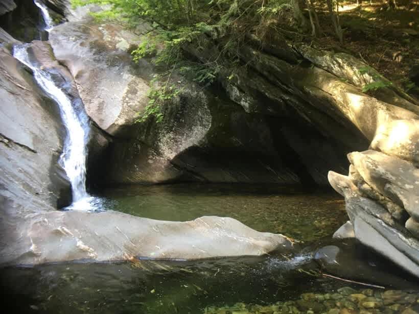 A serene natural waterfall cascades over smooth, grey rock formations into a clear, tranquil pool, surrounded by dense green trees and sun-dappled stones.