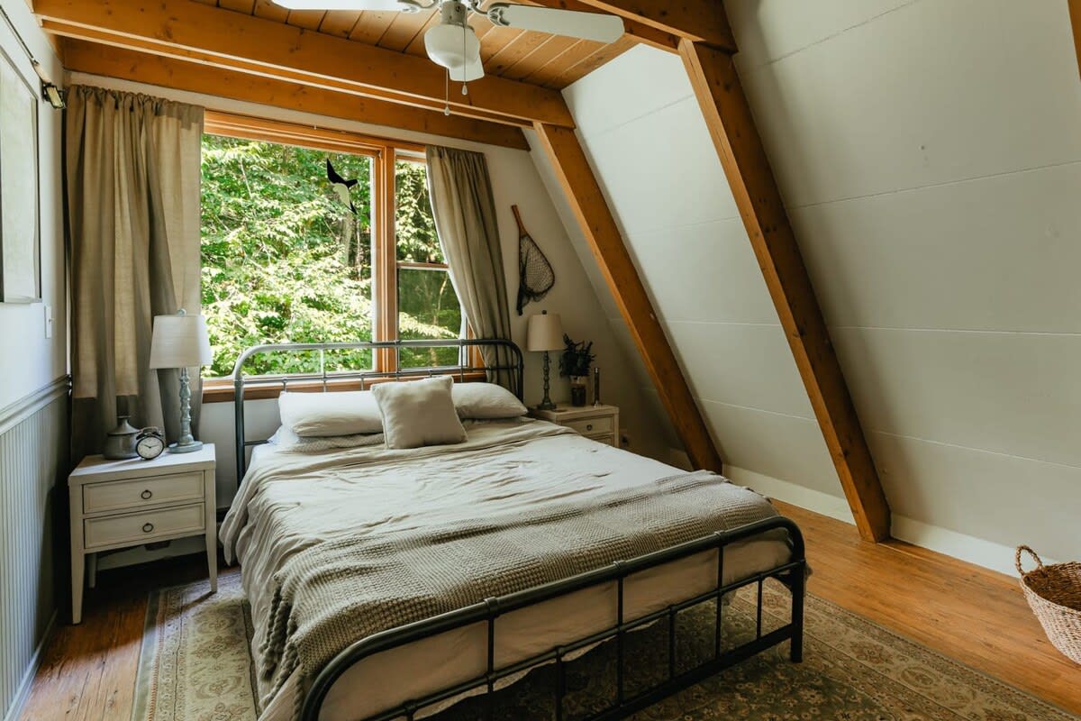 Cozy bedroom with a black metal bed frame, two white nightstands with lamps, and a large window offering a view of lush trees, all under a sloped ceiling with exposed beams.