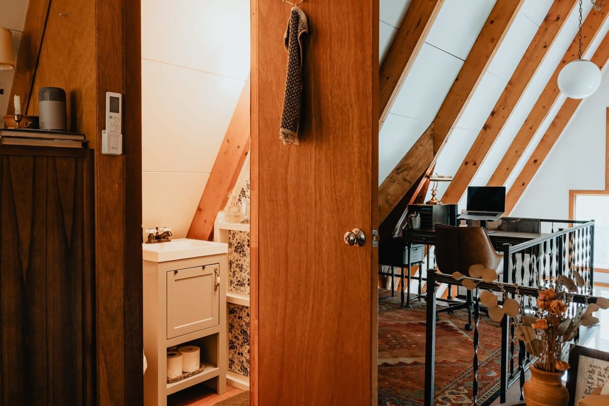 A wooden hallway with a dark metal railing leads to a bathroom with a light gray vanity and patterned wallpaper, all under the sloped ceiling of an A-frame structure.