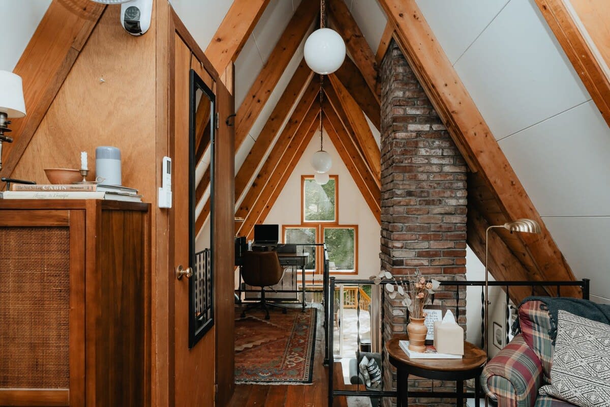 A loft landing overlooks the main living area, featuring a brick chimney, exposed A-frame beams, and a view of the office space with a desk and laptop. A patterned rug is visible on the floor below.
