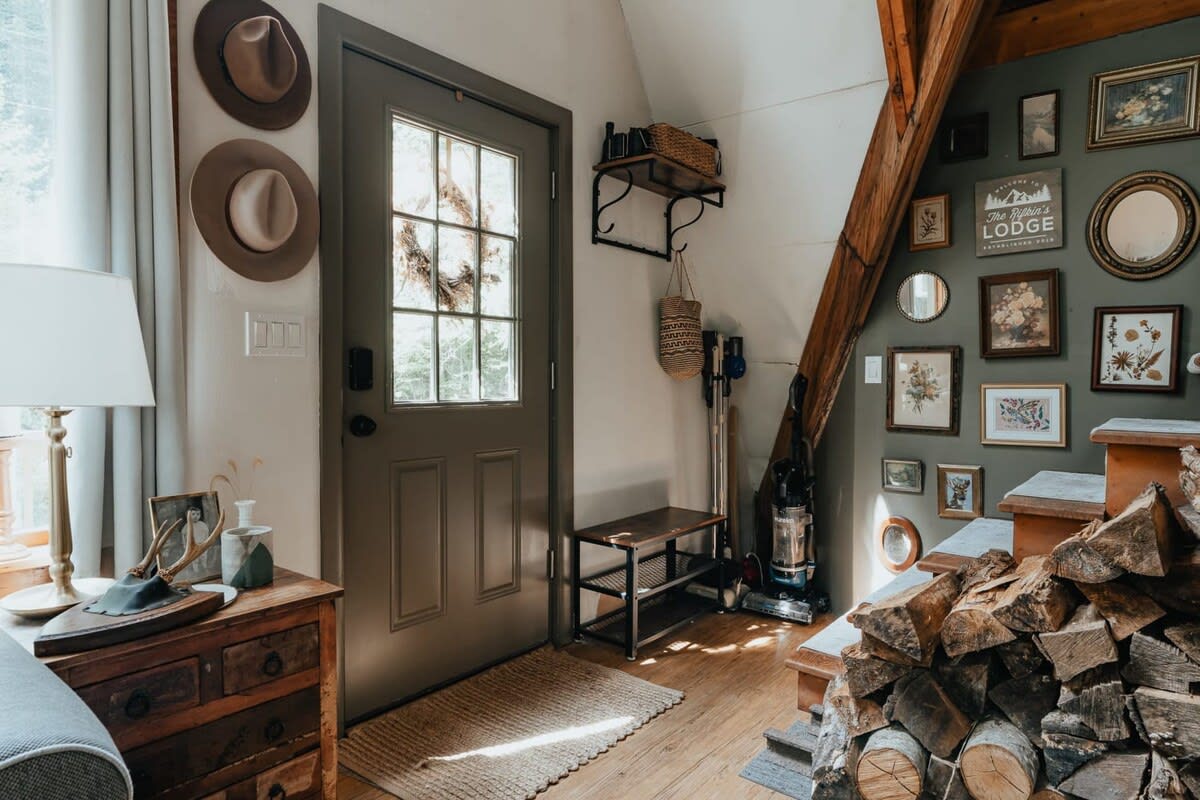 Rustic entryway with a dark green door, decorative hats on the wall, and a neatly stacked pile of firewood beside a wooden staircase, leading to an upper level.