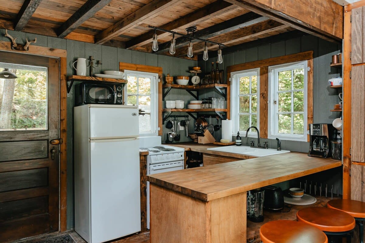 A rustic kitchen with dark green walls and a wood-paneled ceiling, featuring a white refrigerator, a small stove, wooden countertops, open shelving, and a breakfast bar with stools.