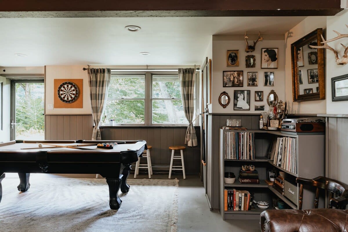 A recreation room features a black pool table, a dartboard on the wall, and a corner shelving unit filled with records and books, all set against wainscoting and large windows.