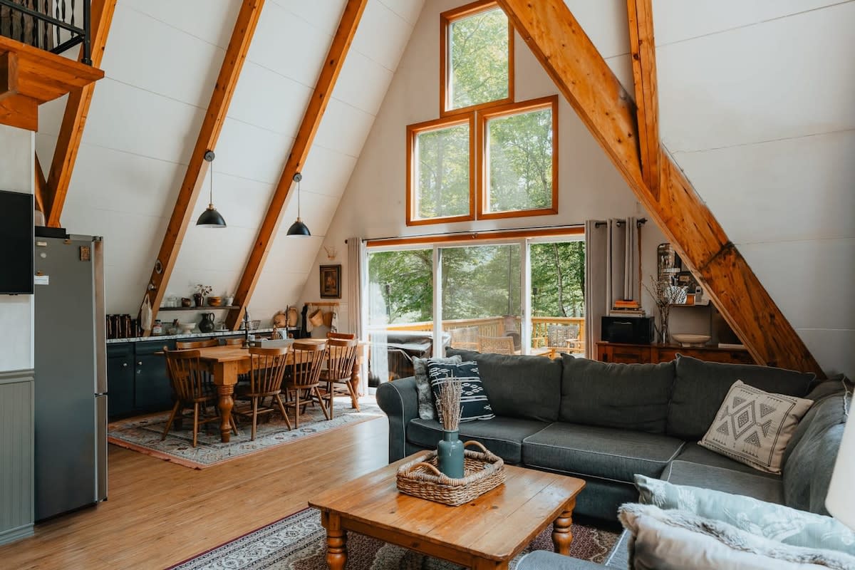 Bright open-concept living and dining area with a gray sectional sofa, a rustic wooden dining table, and large A-frame windows offering views of the surrounding trees.