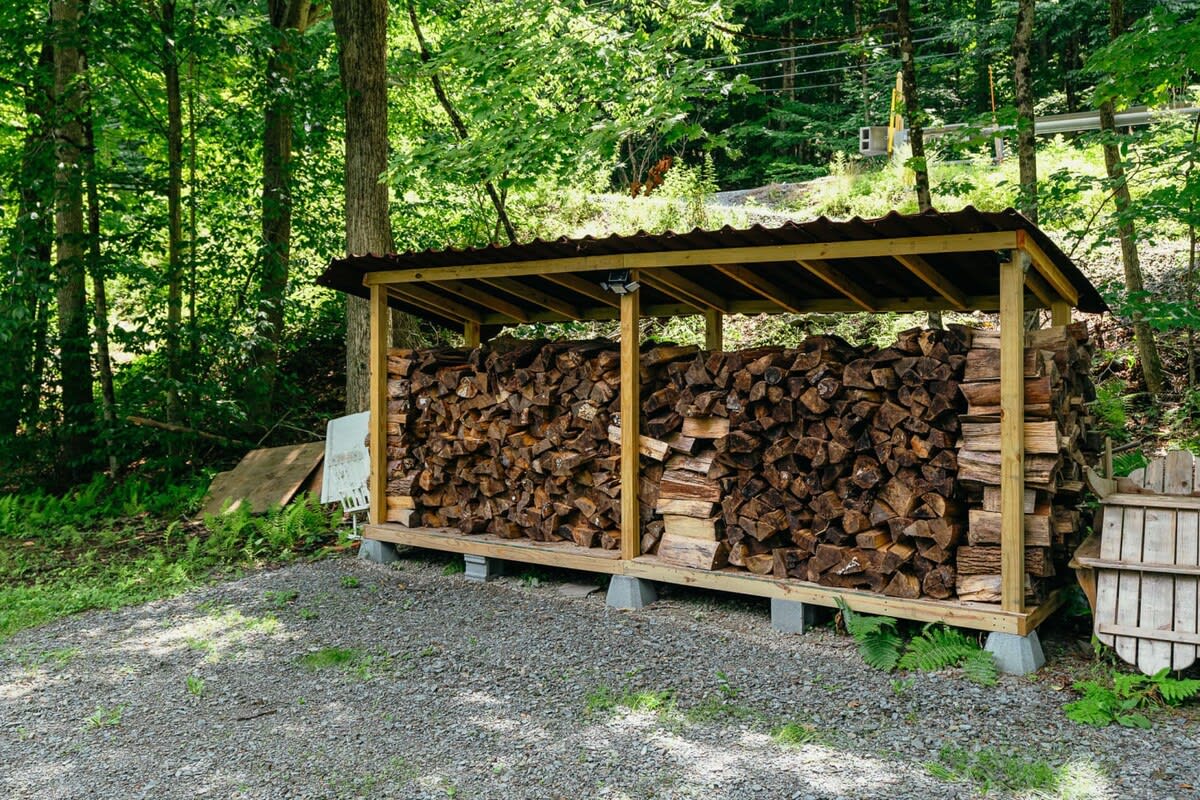 Large, neatly stacked firewood storage shed with a metal roof, located on a gravel patch surrounded by dense green trees. The shed is full of split logs.