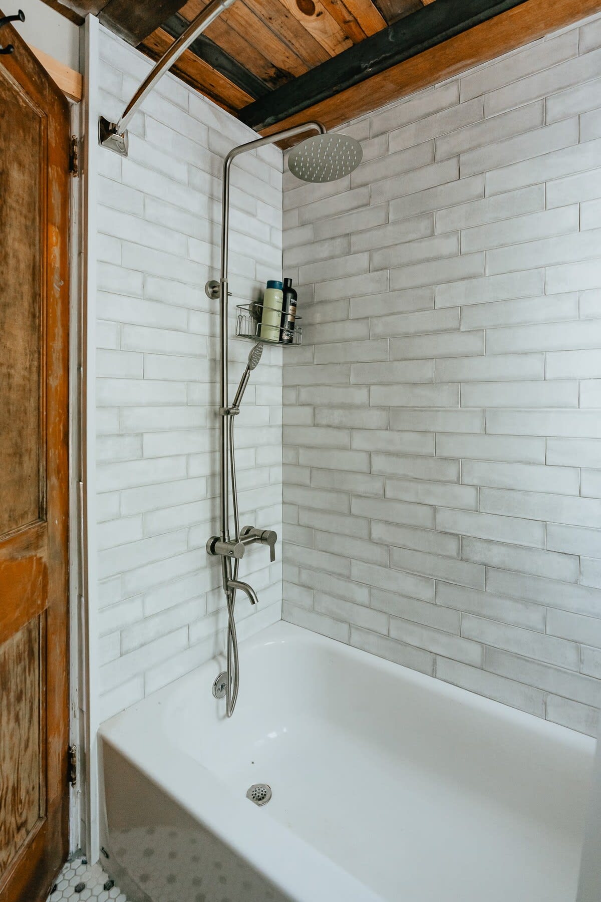 A clean white subway-tiled shower and bathtub area with a modern shower system featuring a rainfall head and handheld sprayer. A rustic wooden door is visible to the left, complementing the cabin aesthetic.