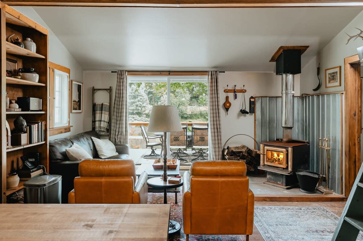 A warm living room featuring a prominent wood-burning stove with a metal flue, two comfortable leather armchairs, a leather sofa, and sliding glass doors leading to an outdoor deck.