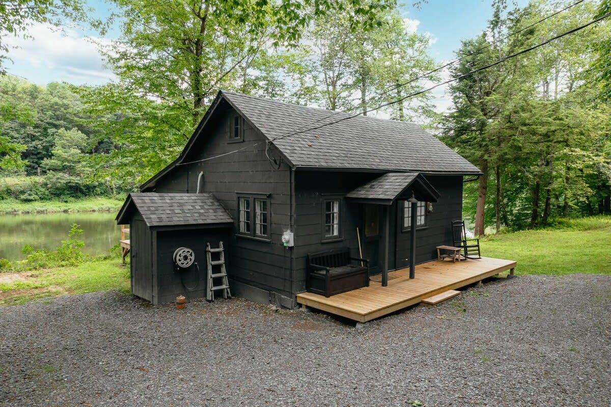 A charming black cabin with a dark shingled roof, featuring a small wooden front porch with a bench and rocking chairs, set against a backdrop of lush trees and a tranquil river.