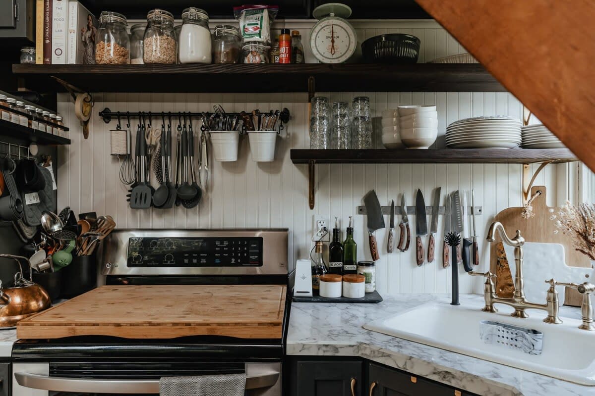 Rustic kitchen area featuring a stainless steel stove with a large wooden cutting board, dark open shelves filled with jars and dishes, and white beadboard backsplash.