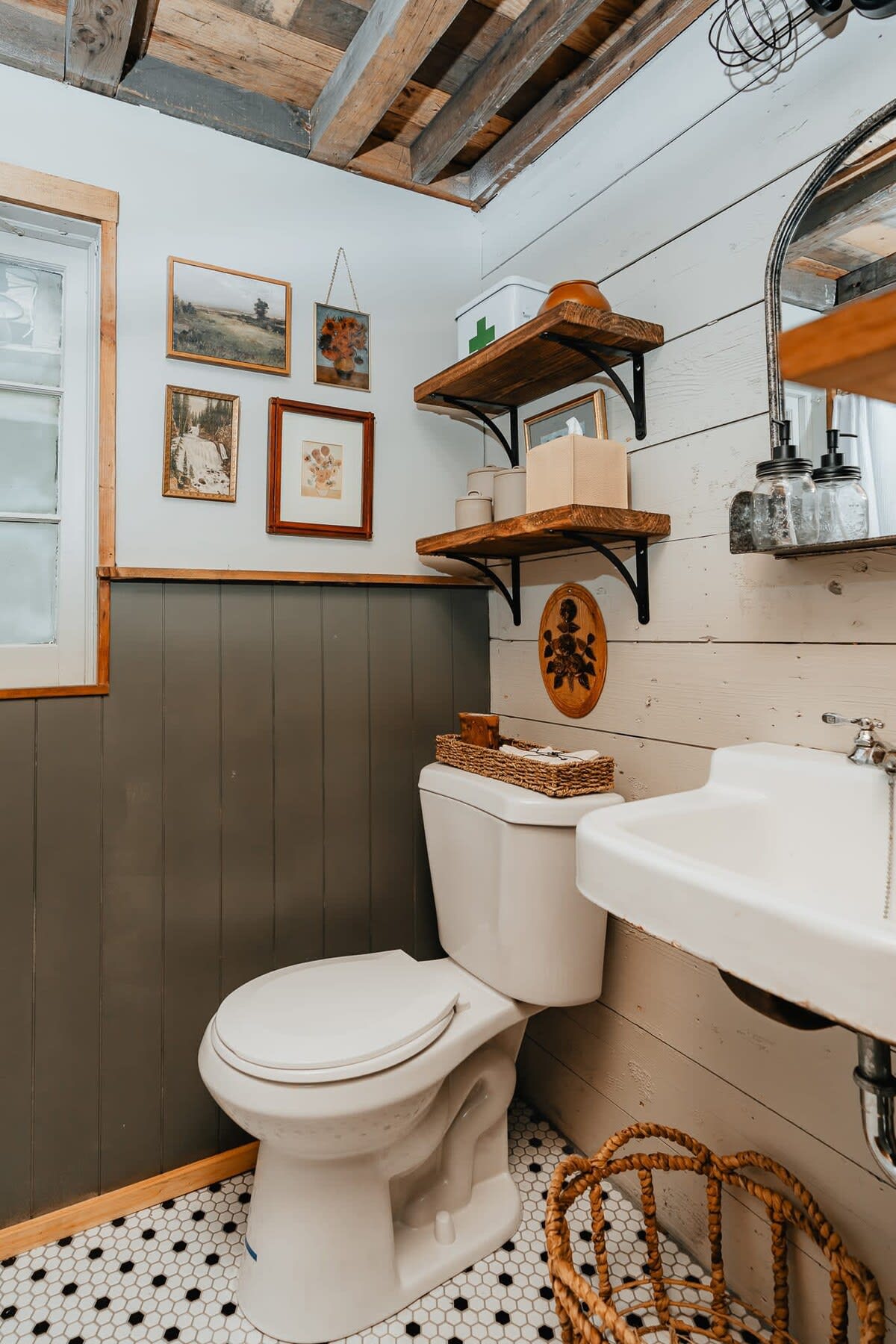 A rustic bathroom with a white toilet and sink, featuring a wainscoting wall painted dark gray below and white shiplap above. Hexagonal black and white floor tiles add a vintage touch, and wooden shelves display decorative items.