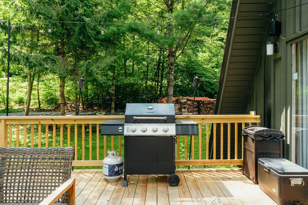 Part of a wooden deck featuring a large black gas grill with a propane tank, and two storage bins, set against a backdrop of green trees. String lights are visible overhead.