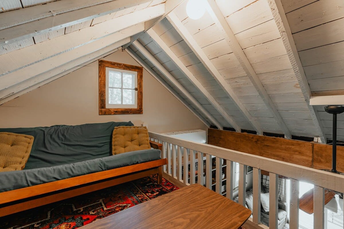 A cozy loft living area with a dark blue futon and a wooden coffee table, situated on a colorful patterned rug. The sloped ceiling with exposed white beams continues the rustic cabin theme, overlooking a lower level.