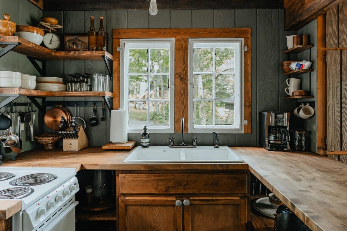 A cozy kitchen corner with a double white sink set into wooden countertops, flanked by two windows, and surrounded by open wooden shelves displaying dishes and kitchenware.