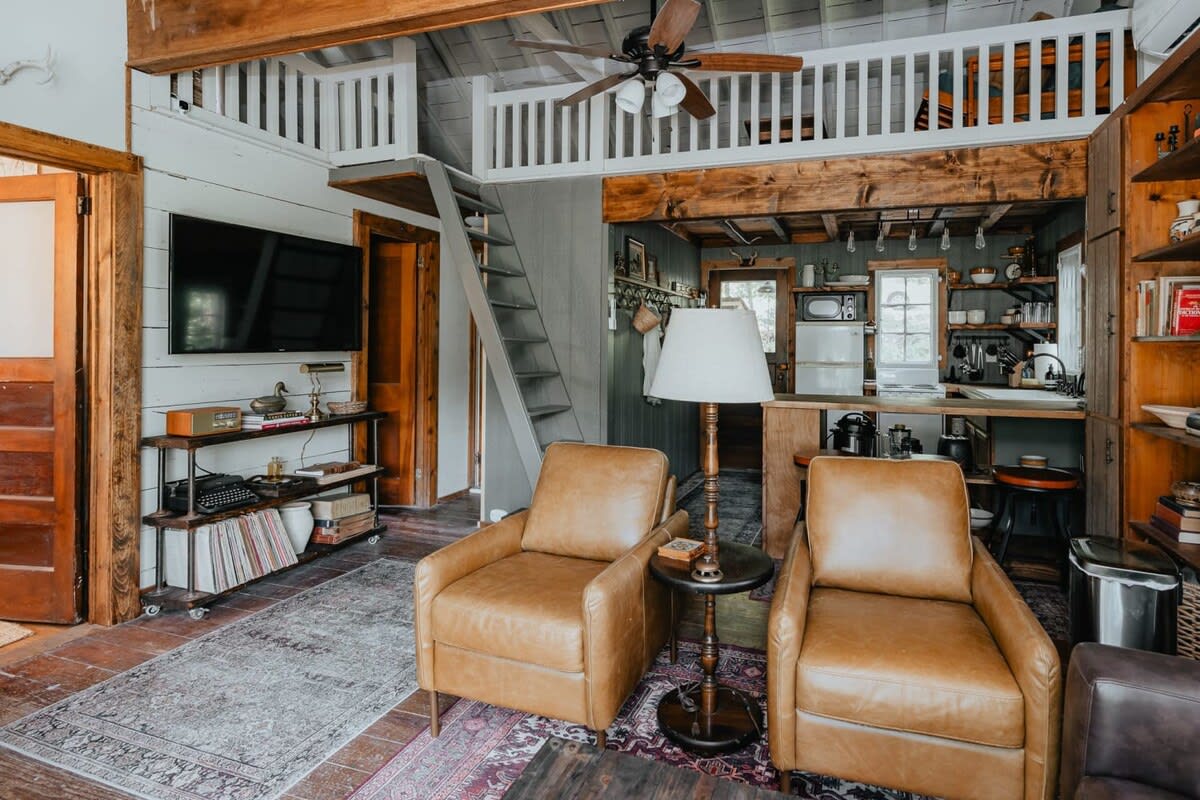 An inviting living space with two tan leather armchairs and a view towards the kitchen and a loft area accessible by a wooden staircase, featuring a ceiling fan and exposed wooden beams.