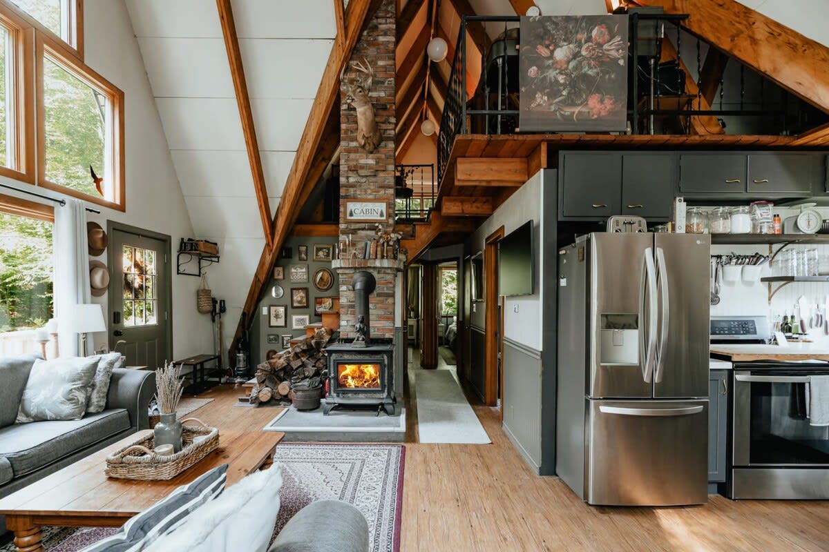 Expansive living area in an A-frame cabin, featuring a wood-burning stove set against a brick fireplace, a gray sectional sofa, and an open-concept kitchen with a large refrigerator.