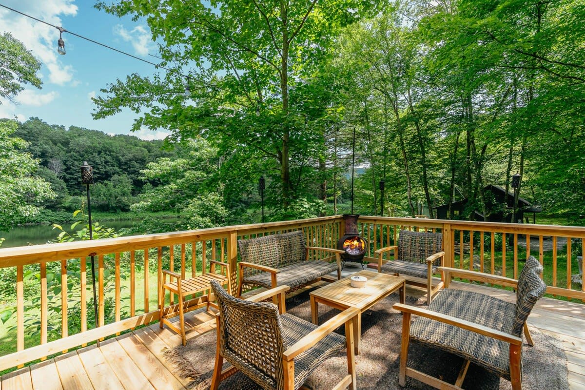 Spacious wooden deck with a full outdoor seating arrangement including a sofa, chairs, and coffee table, centered around a small fire pit. The deck overlooks a lush green forest and a river.