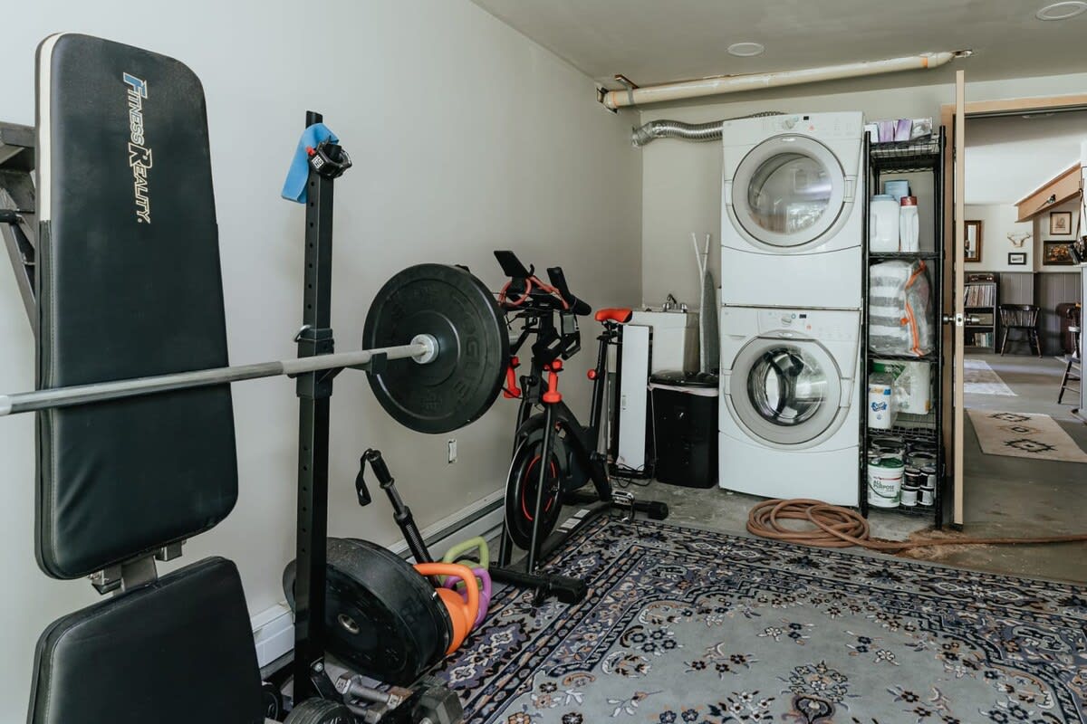 A functional laundry area combined with a home gym, featuring a stacked white washer and dryer, a weight bench, barbell, and exercise bikes on a patterned area rug.