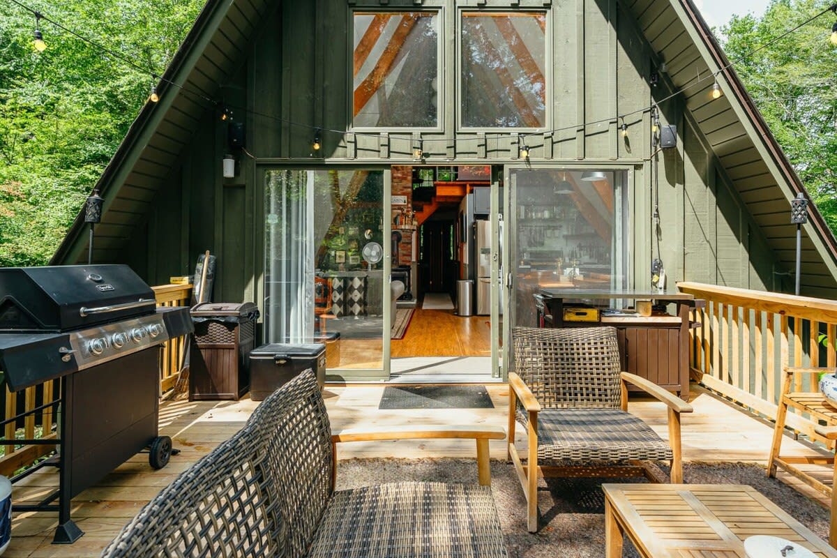 Spacious outdoor wooden deck with a BBQ grill, wicker seating, and string lights, offering a view into the cabin through large sliding glass doors.