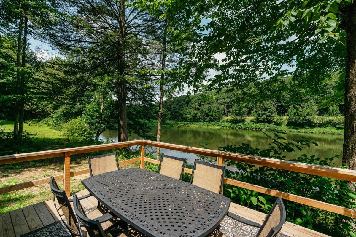 A spacious wooden deck with an outdoor dining set for six, offering scenic views of a calm river surrounded by lush green trees. The deck is enclosed by a wooden railing, perfect for al fresco dining.