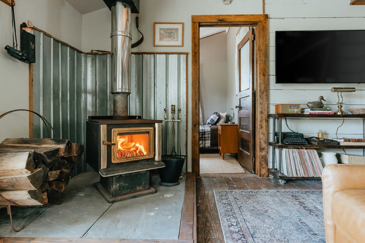 A rustic living area centered around a wood-burning stove with a roaring fire, surrounded by stacked firewood and corrugated metal accents, with a wooden doorway leading to an adjacent room.