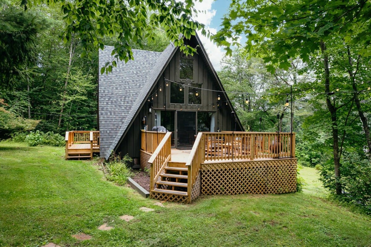 Front view of a dark A-frame cabin with a large wooden deck featuring outdoor seating and string lights, surrounded by a vibrant green lawn and mature trees. A smaller separate deck is visible to the left.