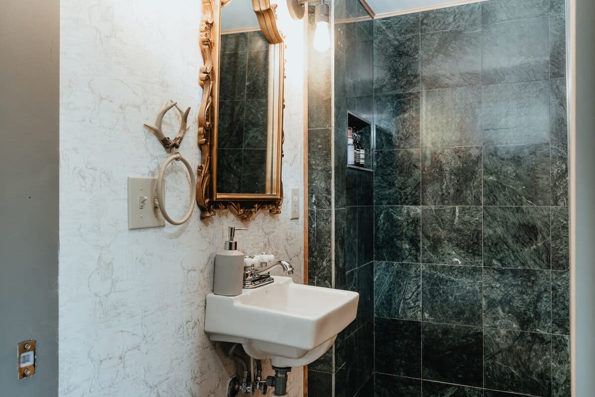 Close-up of a bathroom vanity area featuring a white wall-mounted sink, a decorative gold-framed mirror, and textured light-colored wallpaper, adjacent to a dark green tiled shower.