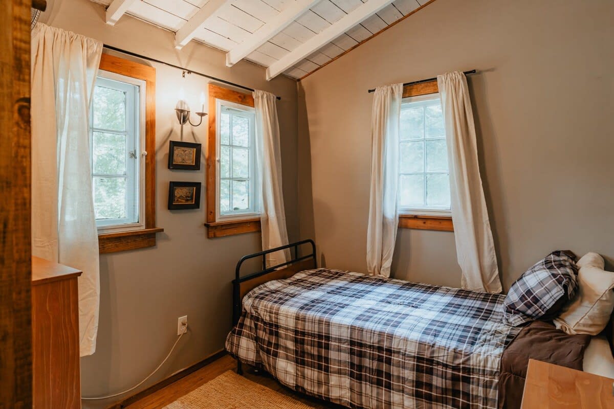 A cozy bedroom with light gray walls and exposed white ceiling beams, featuring a single bed with a plaid comforter, flanked by two windows with simple white curtains and wooden trim.