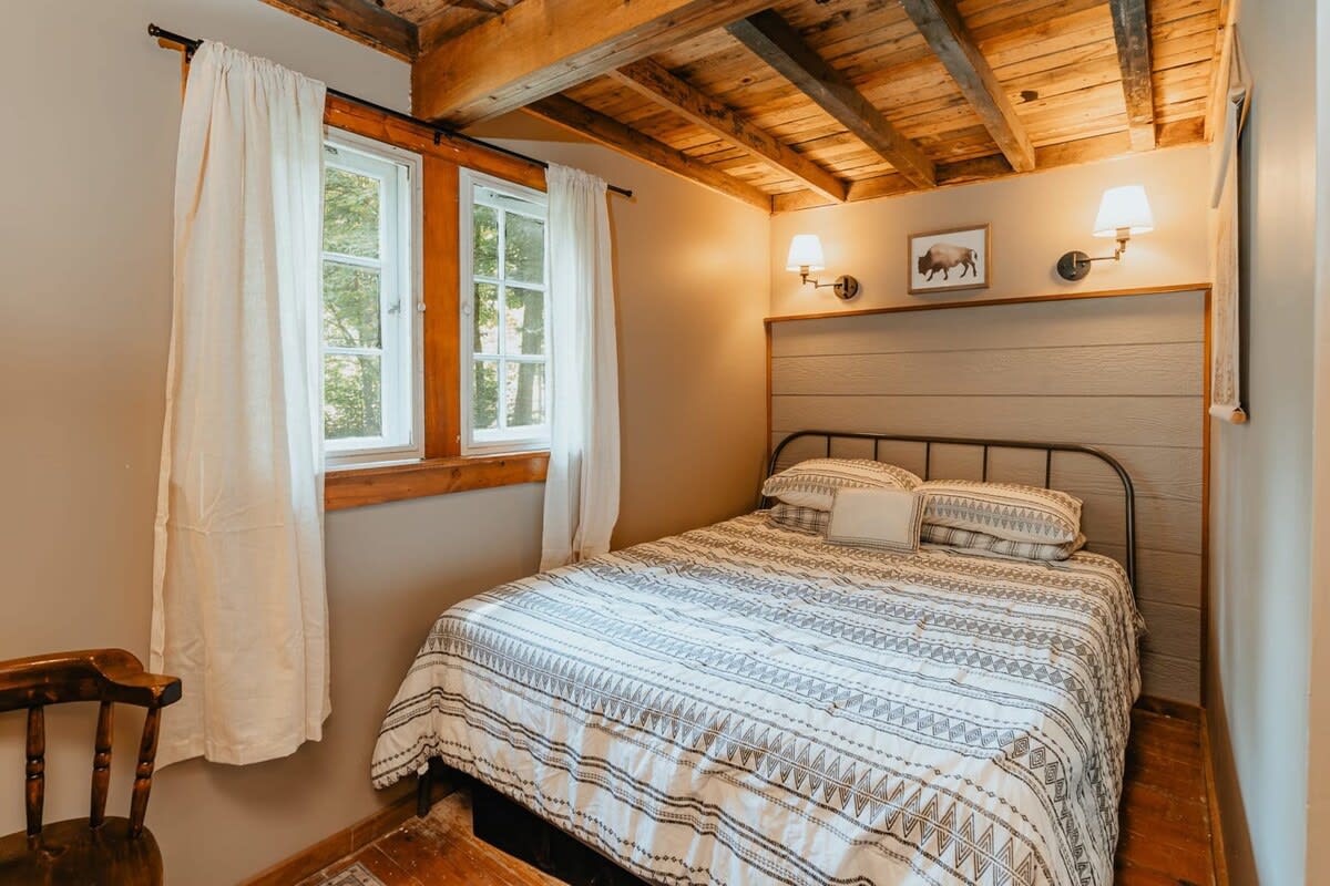 A cozy bedroom with a queen-sized bed featuring a patterned duvet, a slatted wooden headboard, and exposed wooden ceiling beams. Two windows with white curtains overlook greenery, and wall sconces flank a framed bison artwork.